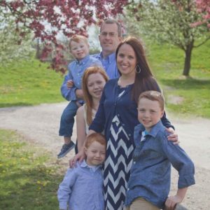 Family photo of the Jacob family. Photo is outdoors with some beautiful greenery and trees that have red leaves. Photo features Jen, her husband, and her four children: Owen (4), Colton (5), Andrew (10), and Katelyn (11).