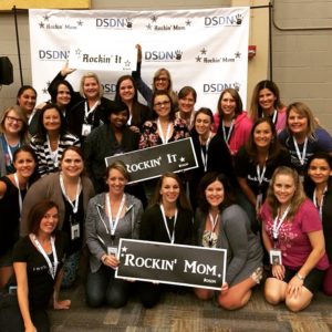 A photo of a group of moms at he last Rockin moms retreat. These moms are huddled up for a group photo in front of a huge DSDN sign. They are also holding Rockin' mom and Rockin' it signs.