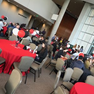 A photo of the gathering room at TEDxACU (Fulks Theater). Red, black and white balloons cover the tables, as well as red table cloths (signature TED colors). People are gathering at the table and breakfast buffet before the big show.