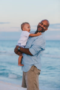 A photo of Vic holding baby Liam on the beach. Liam is touching his beard and Vic is laughing.